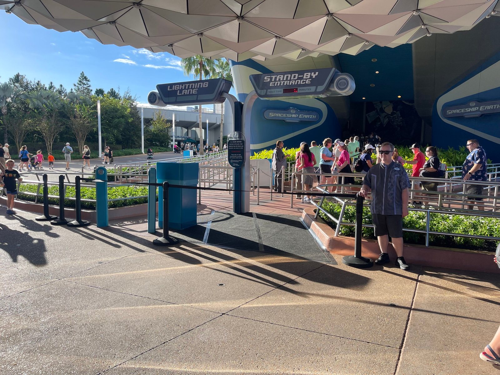 The same Spaceship Earth main entrance closed off with stanchions on a sunny day, with a Cast Member standing beside the barrier and guests in the queue behind