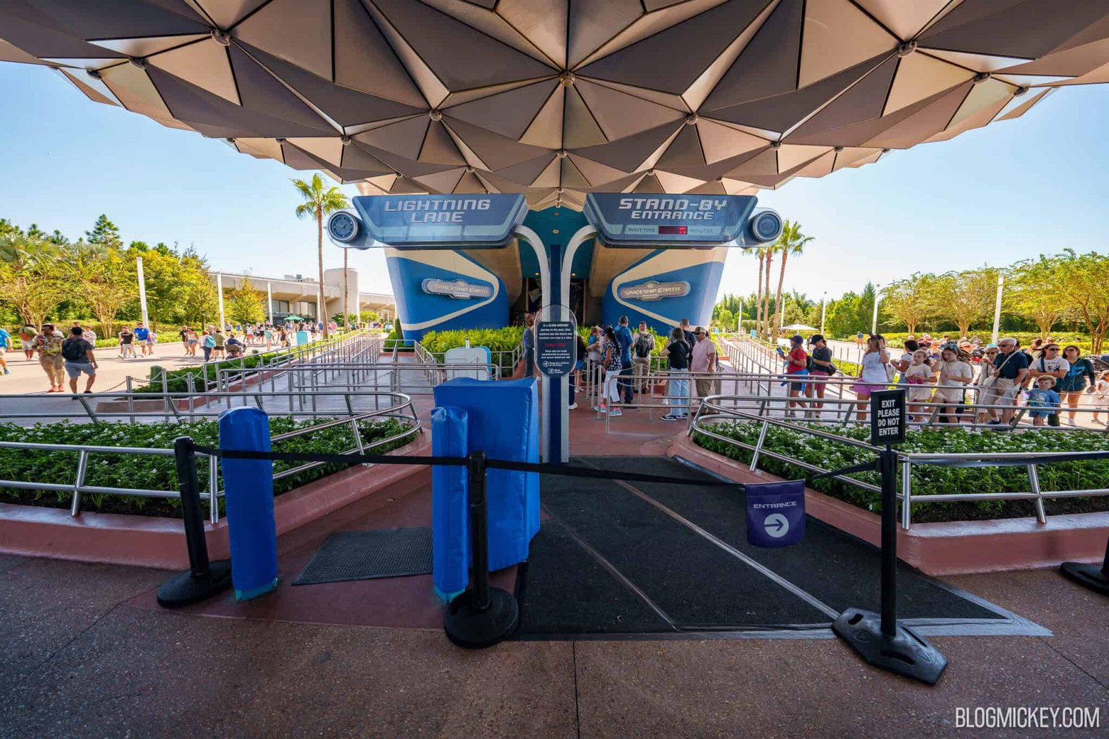 The Spaceship Earth main entrance under the sphere, with a black strap across stanchions, blue padded covers on the posts, an Exit Please Do Not Enter sign and a purple Entrance flag, while guests stand in the queue behind it