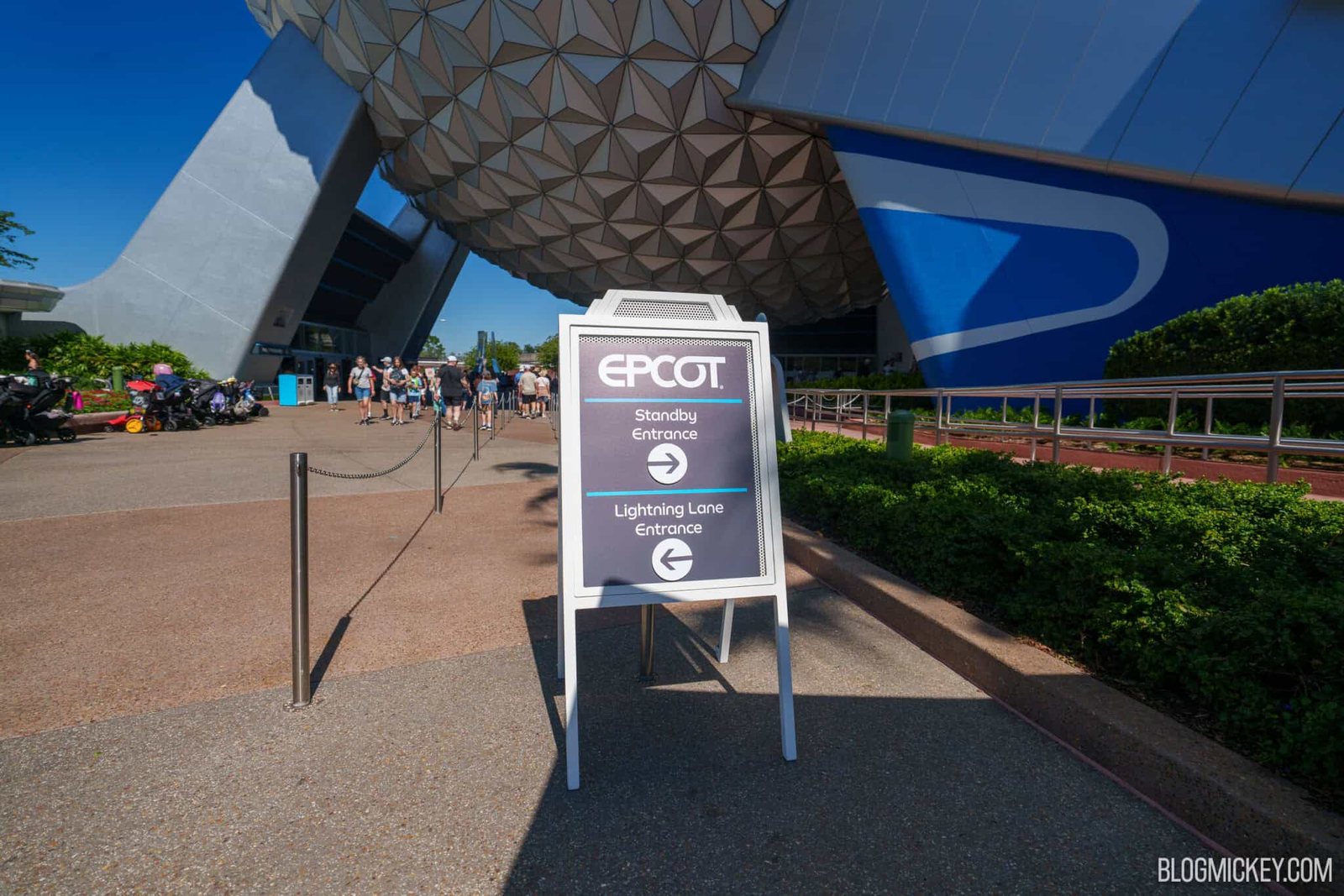 An EPCOT A-frame sign with arrows pointing Standby Entrance to the right and Lightning Lane Entrance to the left, in front of the Spaceship Earth sphere and its support legs
