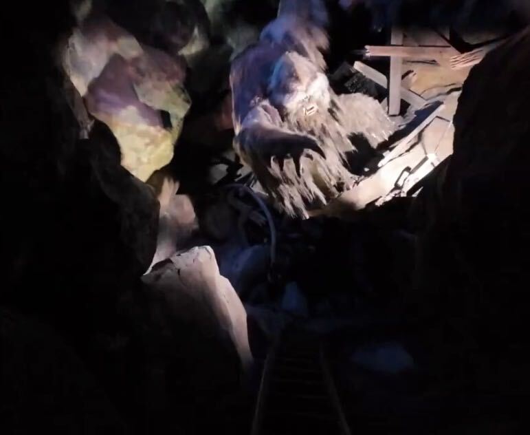 A dim ride-vehicle view of the Expedition Everest Yeti, lit from above with one clawed hand outstretched, beside broken wooden beams, with what appears to be the coaster track in the dark foreground