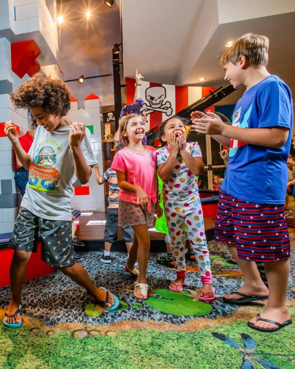 Kids in pajamas and t-shirts dance and laugh on a colorful carpet inside a LEGO-themed hotel lobby, with a skull-and-crossbones banner and LEGO brick walls behind them