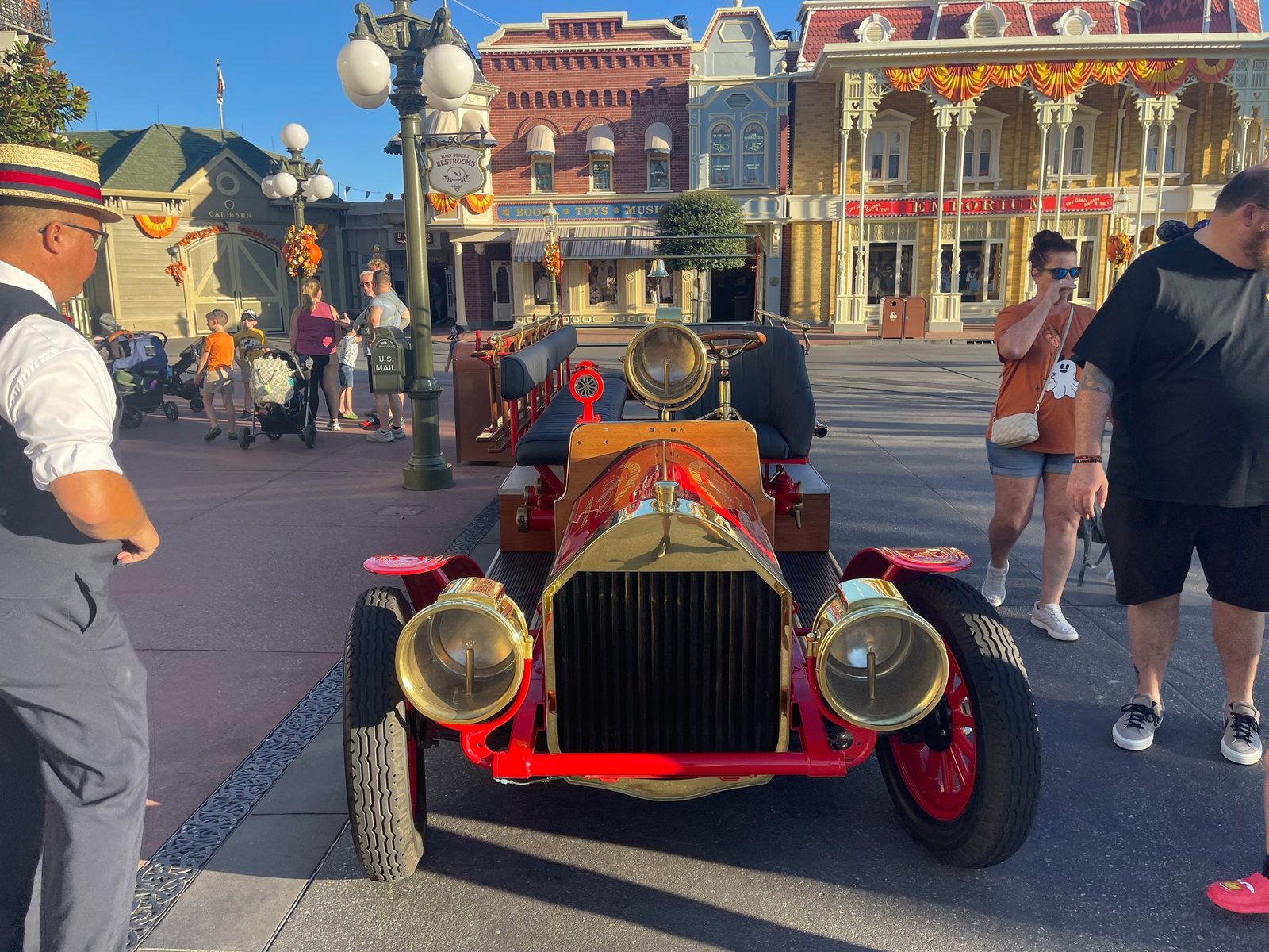 A head-on view of the fire engine at the Town Square curb, showing its brass-framed grille and brass lamps, with a Cast Member in a straw boater standing to the left and guests walking past