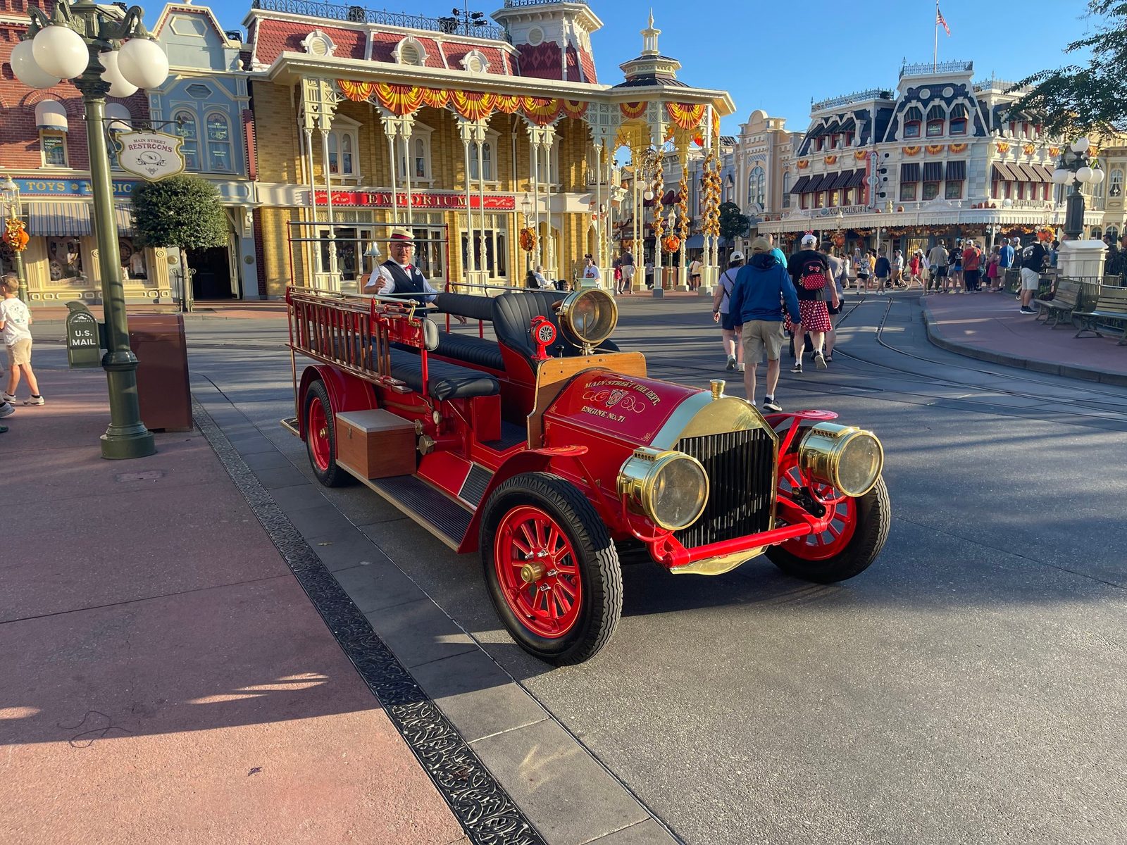 A red open-air fire engine with red-spoked wheels and brass lamps parked at a Town Square curb, with a Cast Member in a straw boater and vest standing aboard at the back ringing a bell, and guests walking past the fall-decorated buildings of Main Street