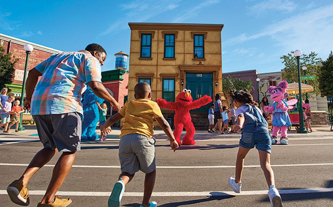 A man and a boy run hand in hand toward Elmo, with a girl running alongside, as Elmo waves his arms in front of a tan brownstone marked 123, with Abby Cadabby and another costumed character nearby in Sesame Street Land