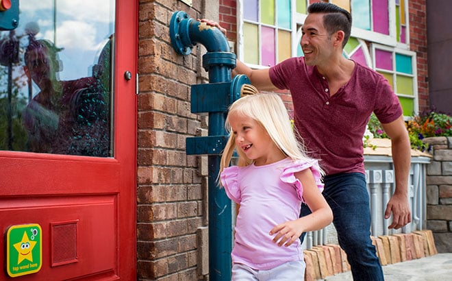 A smiling young girl in a pink top runs past a red door and a blue pipe on a brick wall in Sesame Street Land, with a man laughing behind her