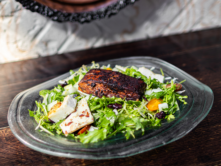 A blackened halibut fillet on a bed of arugula and frisée with feta brûlée, tomatoes and dried cranberries on a glass plate