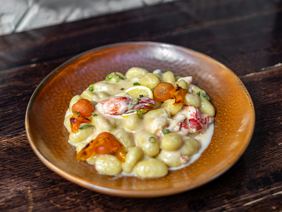 A bowl of gnocchi in a creamy white sauce with lobster, a lemon slice, blistered tomatoes and chopped herbs