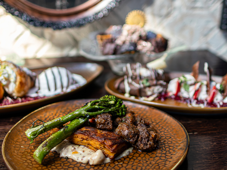 A spread of Toothsome National Chocolate Day dishes on a wooden table, with beef bourguignon and broccolini in front and the pistachio croissant, the corn and chocolate dessert and the chocolate sundae behind it