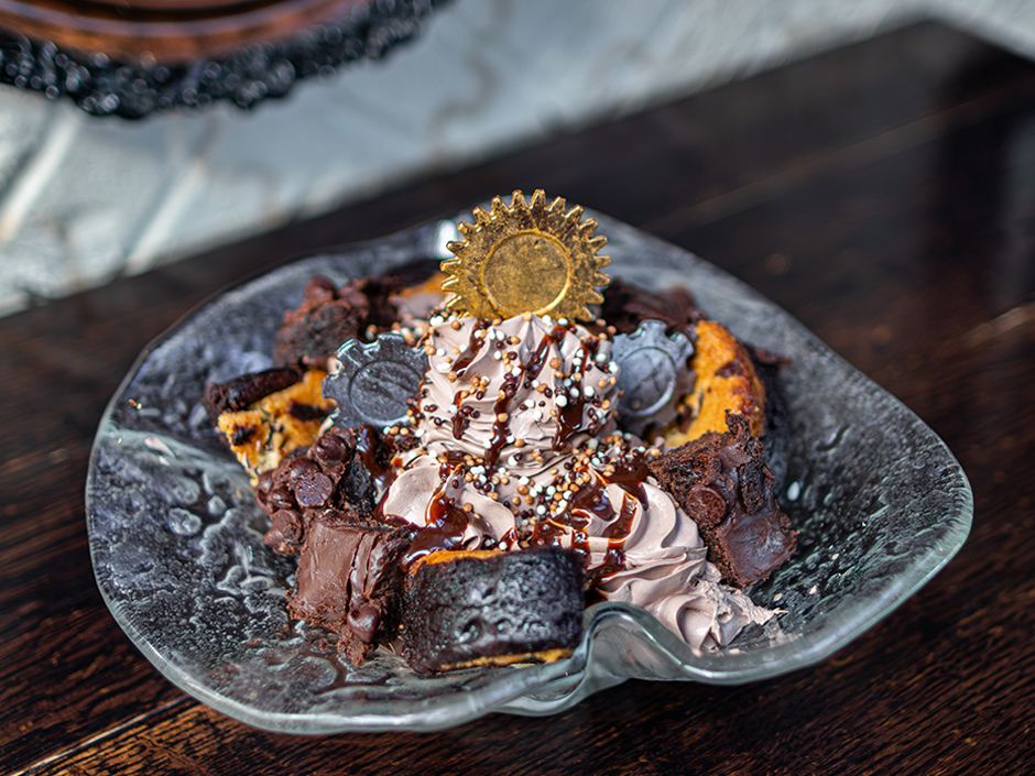 A chocolate sundae on a glass plate piled with brownies, cookie pieces, chocolate whipped topping, sprinkles and a gold chocolate gear on top