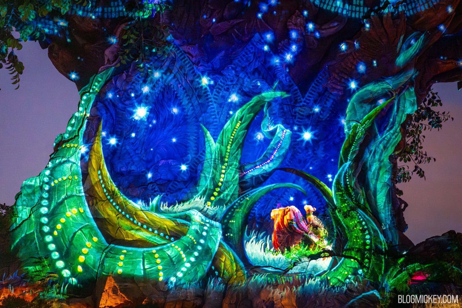 A close-up of the Tree of Life trunk covered in glowing projections of a blue starry sky, green grass blades and dotted vines, with a glowing adult lion and a smaller lion at the lower right