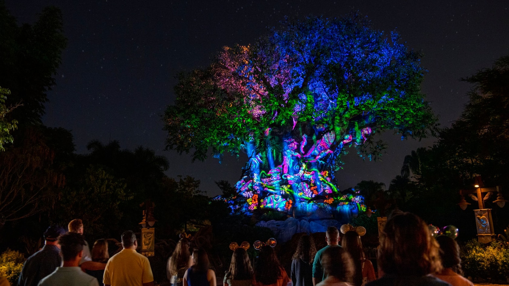 Guests silhouetted in the foreground watch the Tree of Life at night, its branches lit blue, green and pink and its trunk covered in glowing projected animals and flowers