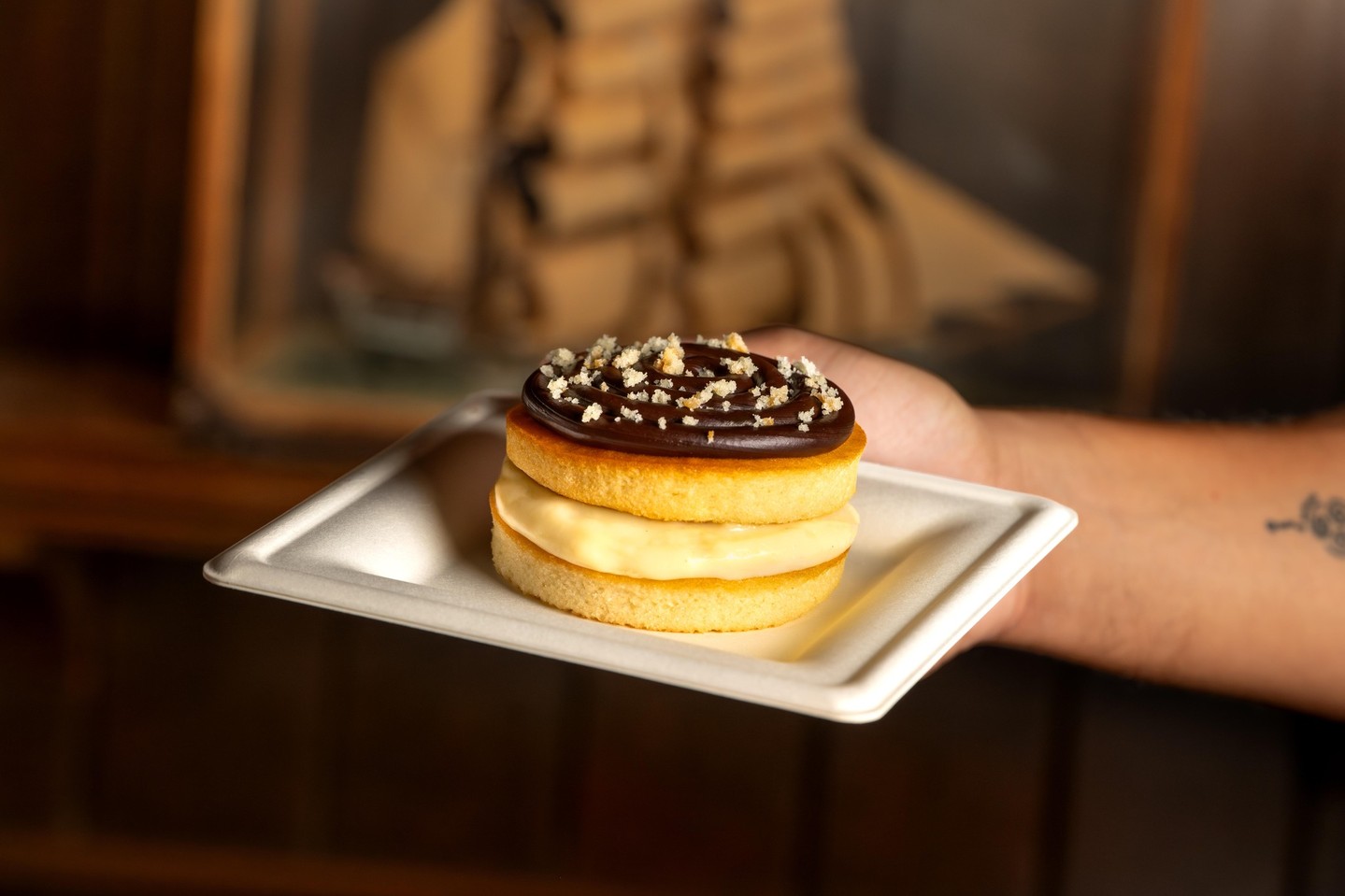 A small round Boston cream pie on a rectangular molded fiber plate, two layers of yellow cake around pale cream, topped with a spiral of dark chocolate ganache and cake crumbs