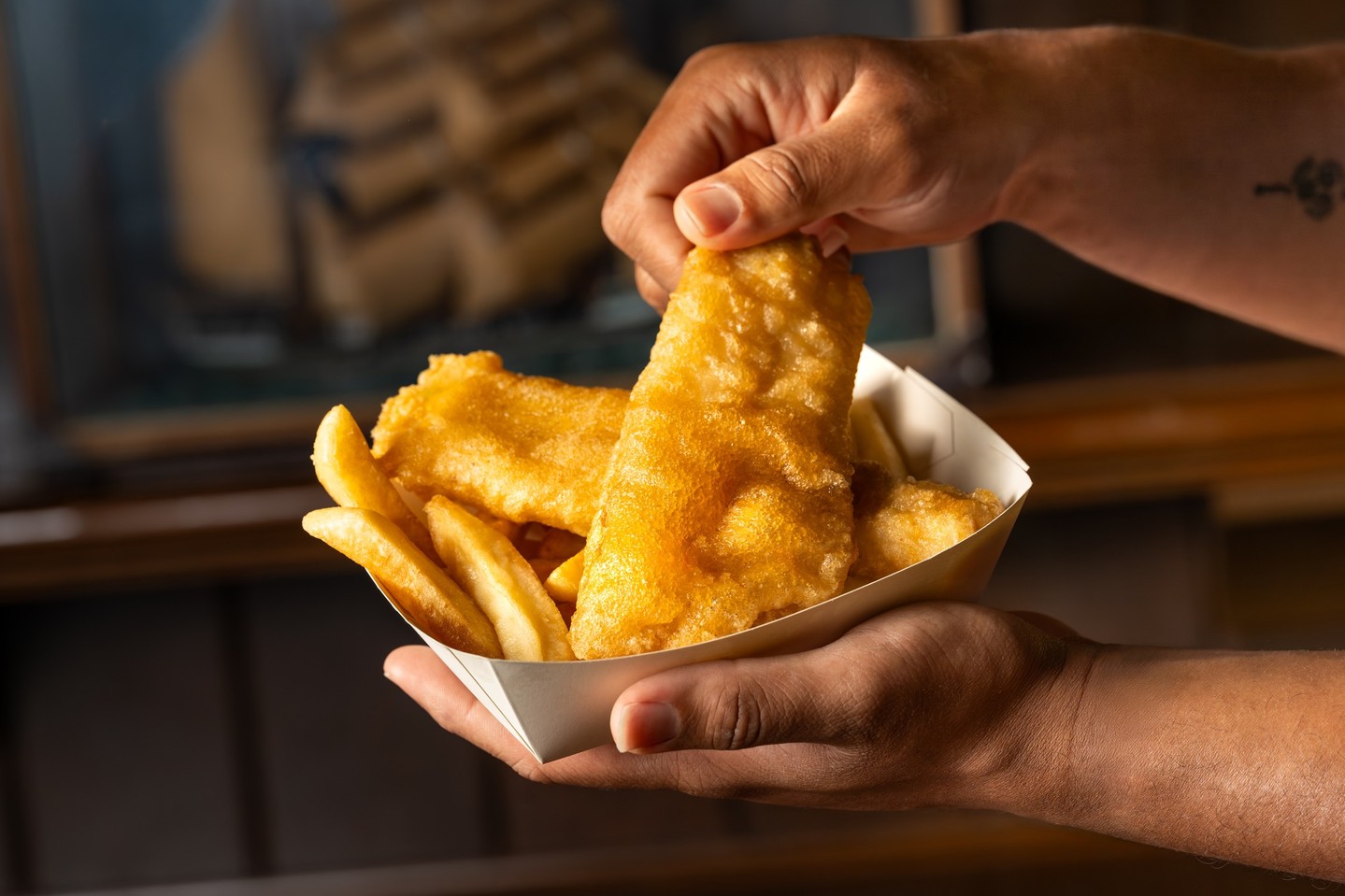 A hand lifts a long golden battered fish fillet out of a white paper tray of fish and French fries, with a model sailing ship blurred in the background