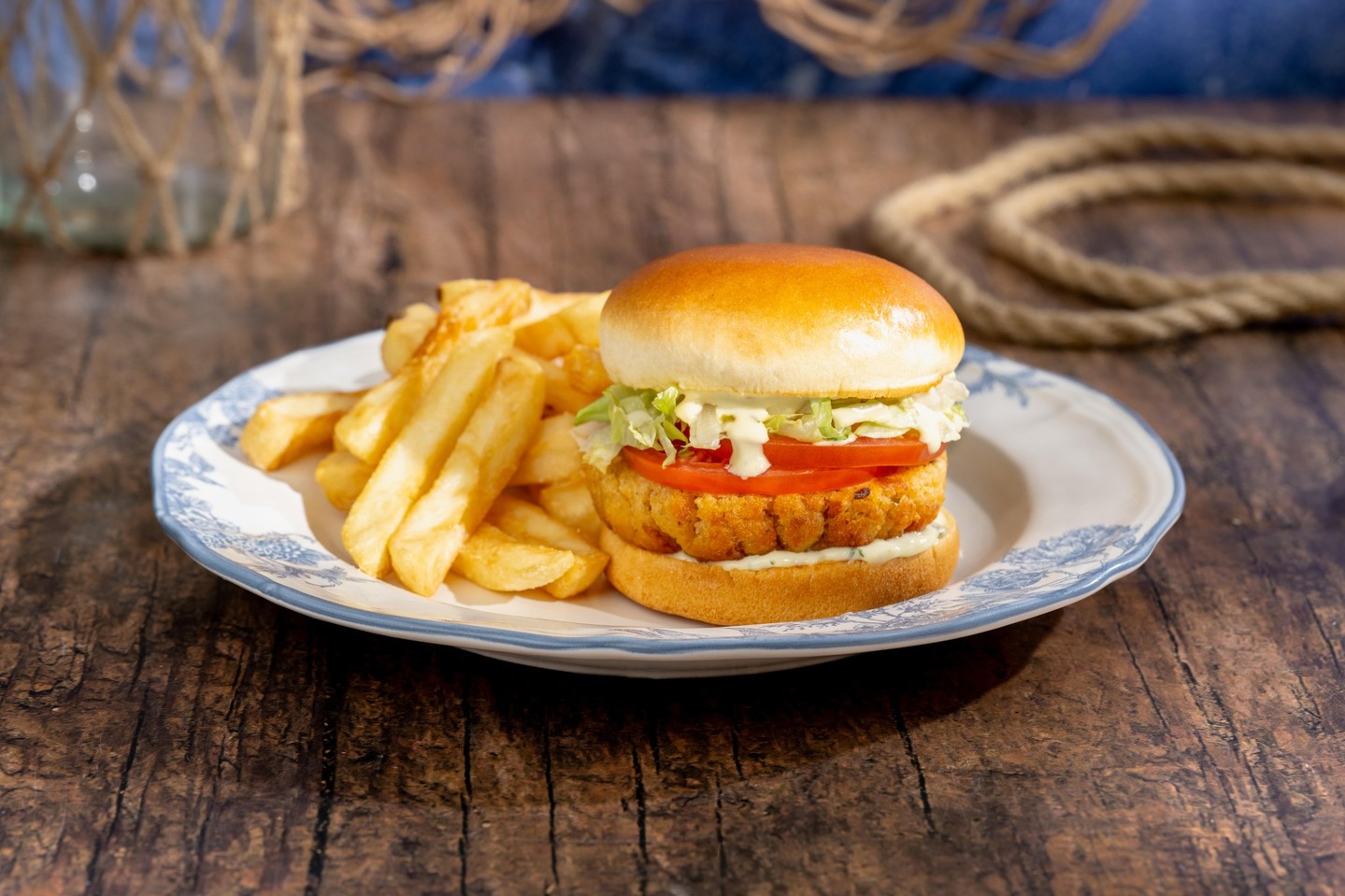 A plant-based crab cake sandwich on a glossy toasted bun with tomato, shredded lettuce and a creamy sauce, next to thick-cut fries on a blue-rimmed plate, with rope and fishing net in the background