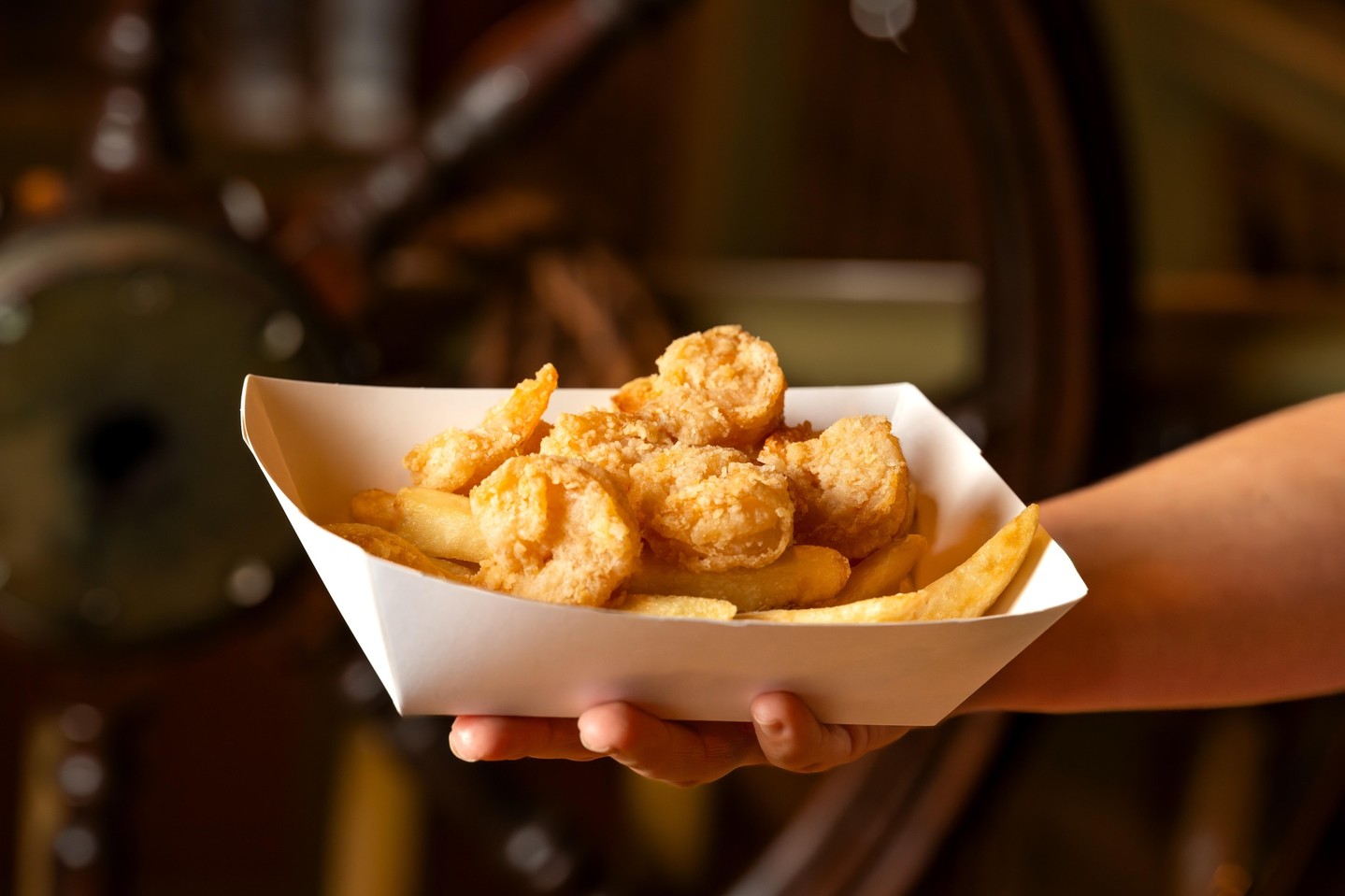 An outstretched arm holds a white paper tray of breaded fried shrimp on top of French fries, in front of a blurred wooden ship wheel