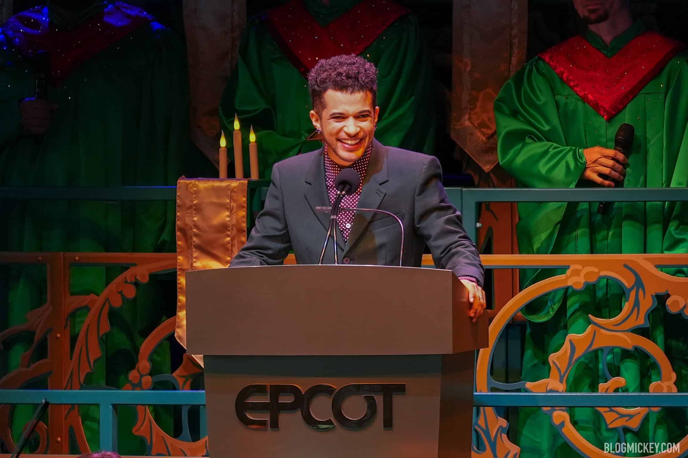 Jordan Fisher, in a dark suit and patterned shirt, smiles from behind a podium marked EPCOT, with candles and choir members in green and red robes behind him