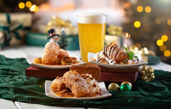 Three plates of beignets, two on a wooden stand: one with caramelized bananas, one drizzled with chocolate and topped with marshmallows, and one dusted with cookie crumbs beside a Biscoff cookie, with a pint of golden beer behind them