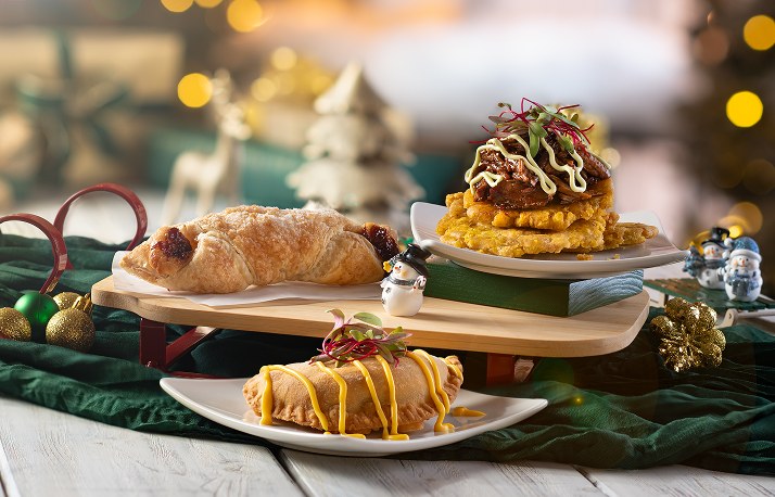 Shredded beef topped with a creamy drizzle and microgreens on a stack of golden tostones, next to a flaky pastry and an empanada drizzled with yellow sauce, on wooden serving boards with snowman figurines