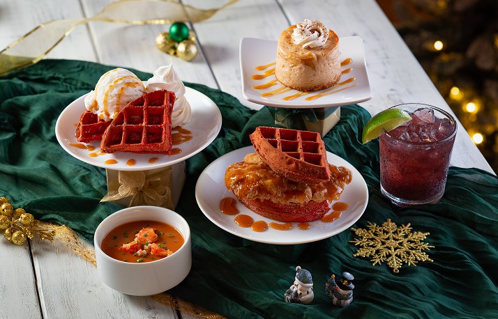 Red waffles with ice cream and caramel, a red waffle stacked on fried chicken, a cup of orange lobster bisque with lobster pieces, a small cheesecake and a purple iced drink with a lime wedge, on a green cloth with holiday decorations