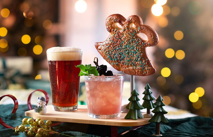 A large bell-shaped soft pretzel with blue, green and red sprinkles stands behind a pint of amber beer and a pink cocktail garnished with blackberries and mint, on a small wooden stand with miniature evergreen trees