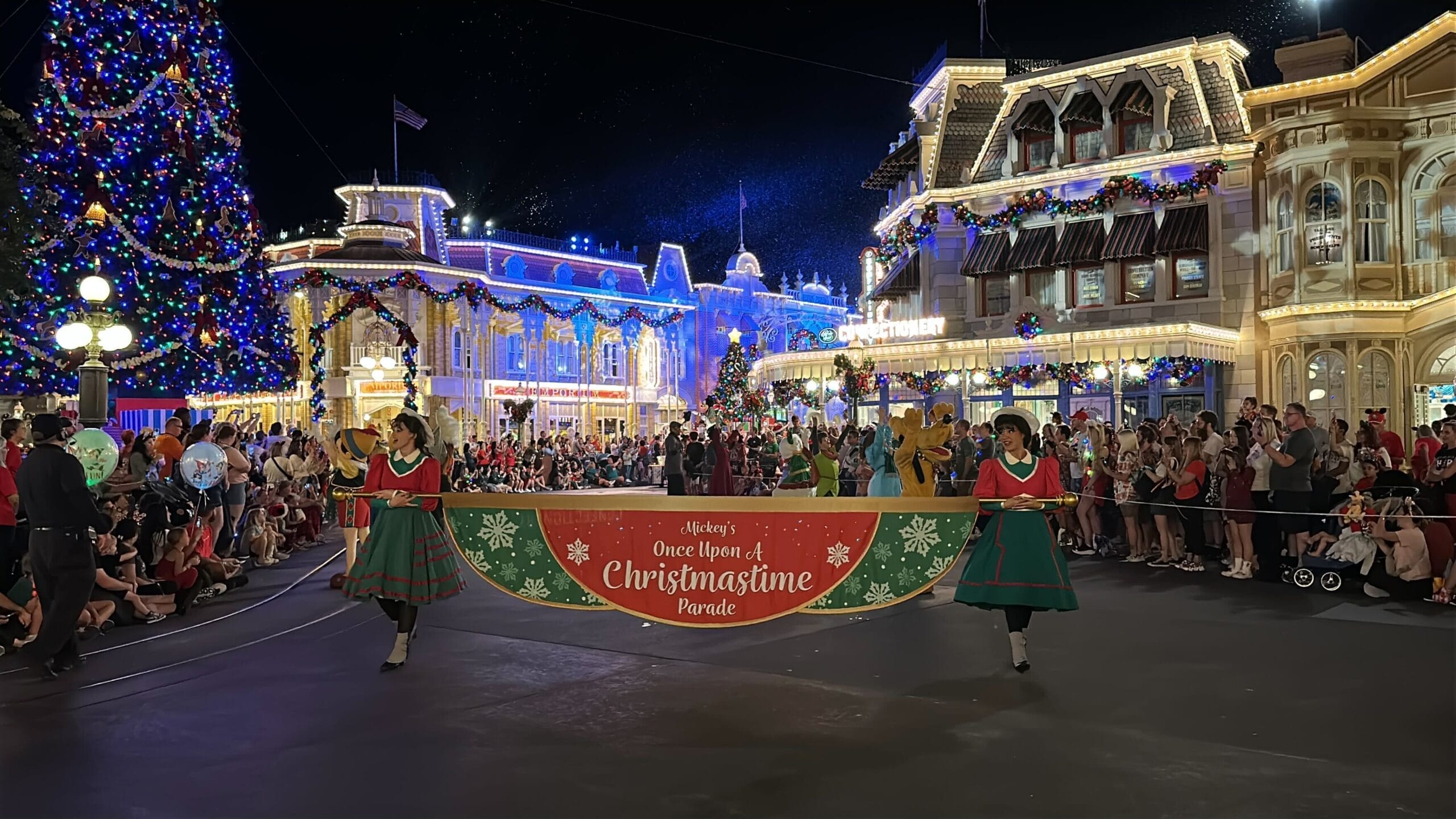 Two performers in red and green dresses carry a banner reading Mickey's Once Upon A Christmastime Parade down Main Street, U.S.A. at night, with a giant lit Christmas tree on the left, garland-draped buildings and crowds lining the curb