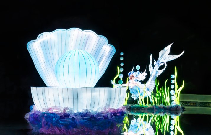 A giant glowing white and pale blue clam shell lantern holding a luminous pearl, beside a mermaid lantern with a white flowing tail surrounded by bright green seaweed lanterns, reflected in dark water