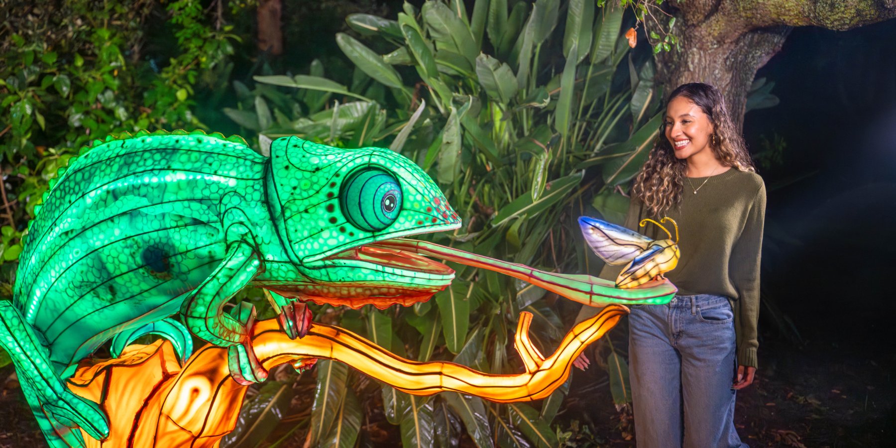 A large glowing green chameleon lantern perched on an orange lit branch, its long tongue stretched toward a glowing winged insect lantern, as a smiling woman in an olive sweater and jeans looks on among dark tropical foliage