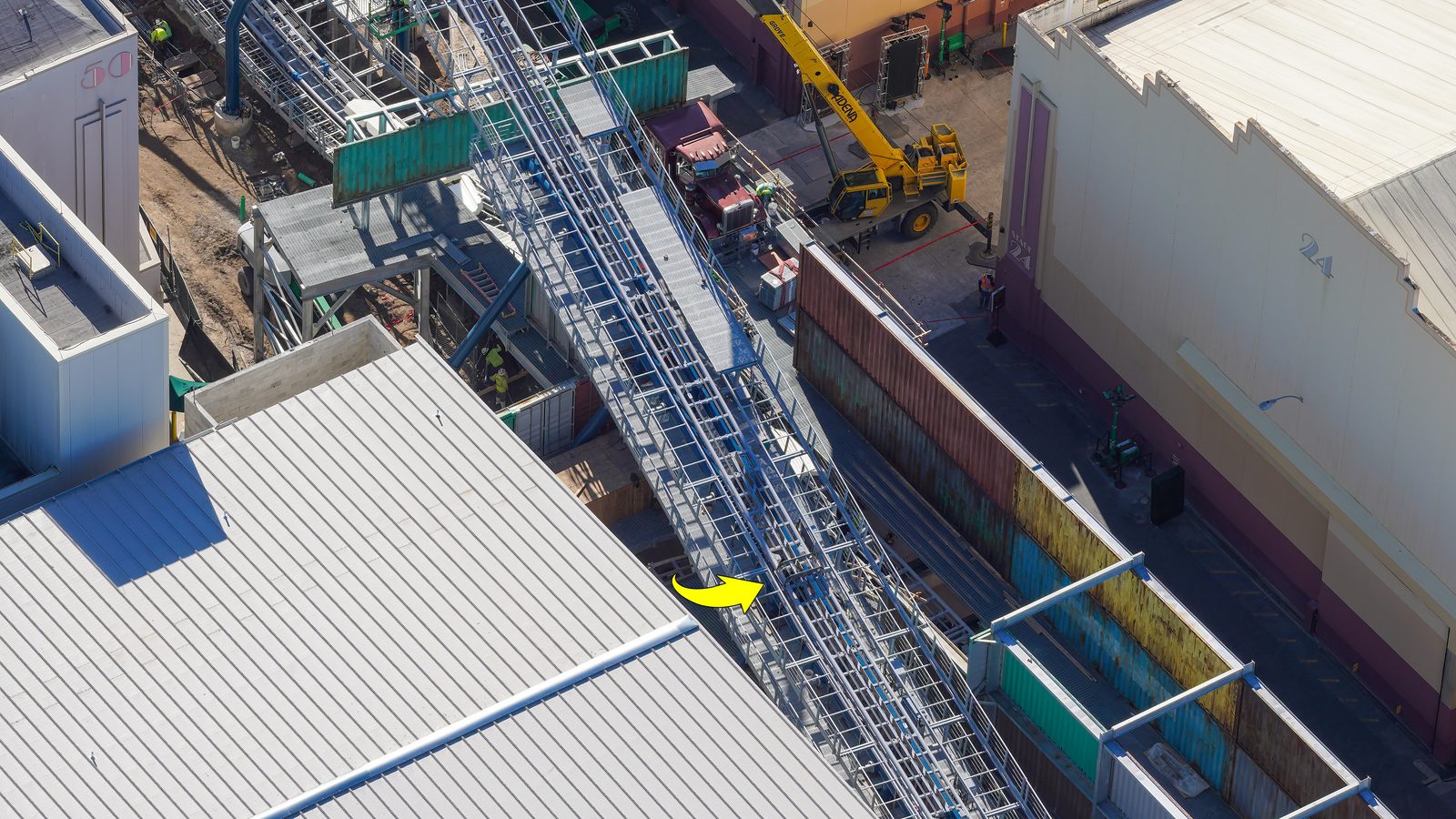 Aerial view of a long gray steel brake run with catwalks on both sides, a yellow arrow pointing at a spot on the track, weathered red, yellow and teal shipping container walls running alongside it, a yellow crane and a dark red truck cab beyond the walls, and a large white show building roof in the foreground
