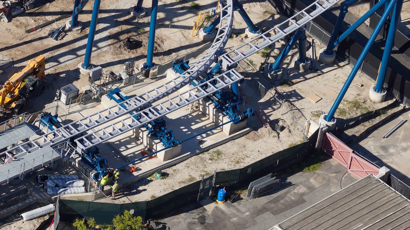 Aerial view of two gray coaster track sections, one curved and one straight, mounted on a concrete pad with blue actuator machinery beneath them, blue support columns around the pad and workers in high-visibility vests standing at one end