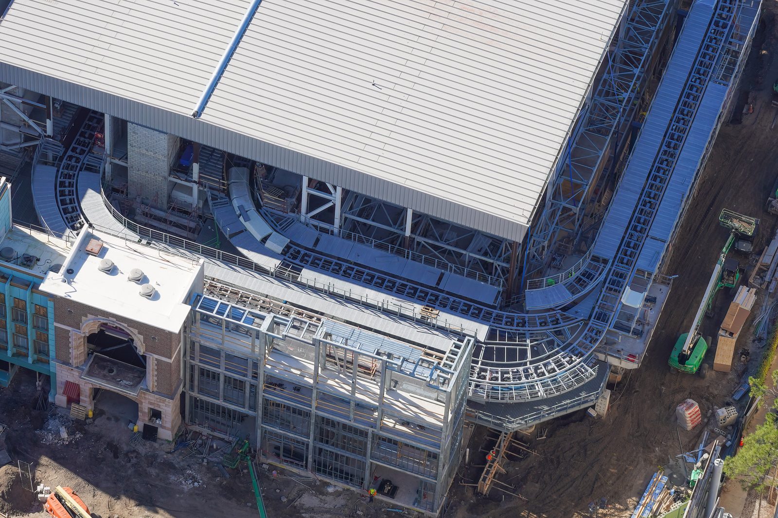 Aerial view of a large white-roofed show building with gray coaster track curving in a U shape along its open side, exposed steel framing and catwalks, a brick facade with an arched entrance at lower left and a green lift on the dirt at right