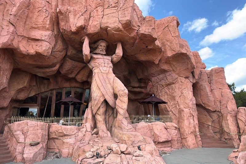 The rockwork facade of Mythos Restaurant, with a giant carved statue of a bearded figure holding up the rock in front of an outdoor patio with umbrellas and arched windows