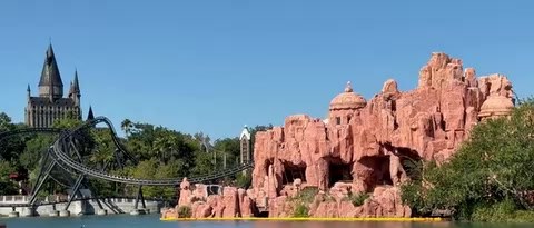A wide view across the Islands of Adventure lagoon of the pink rockwork and domes of Mythos, with Hogwarts castle and the dark track of Hagrid's Magical Creatures Motorbike Adventure to the left and a yellow barrier floating along the base of the rocks