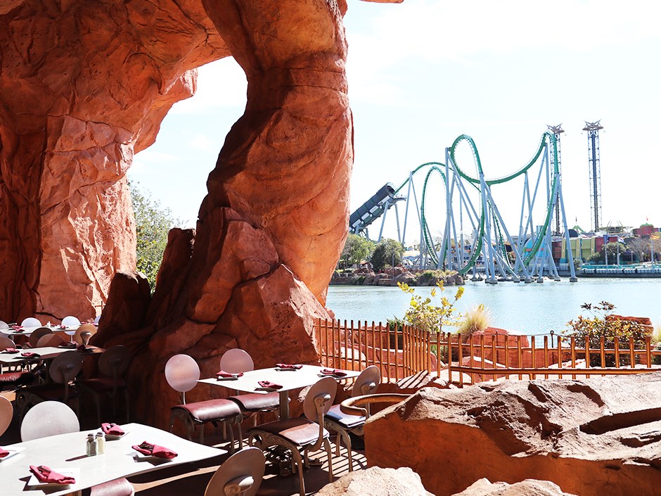 Outdoor tables at Mythos set under a rock arch, looking out across the lagoon to the green Incredible Hulk Coaster