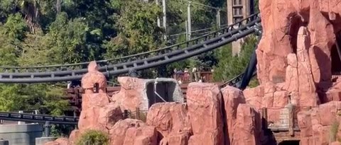 Close view across the water of the craggy pink rockwork behind Mythos, where an opening cut into the rock shows grey framework inside, with the dark track of Hagrid's Magical Creatures Motorbike Adventure running behind it