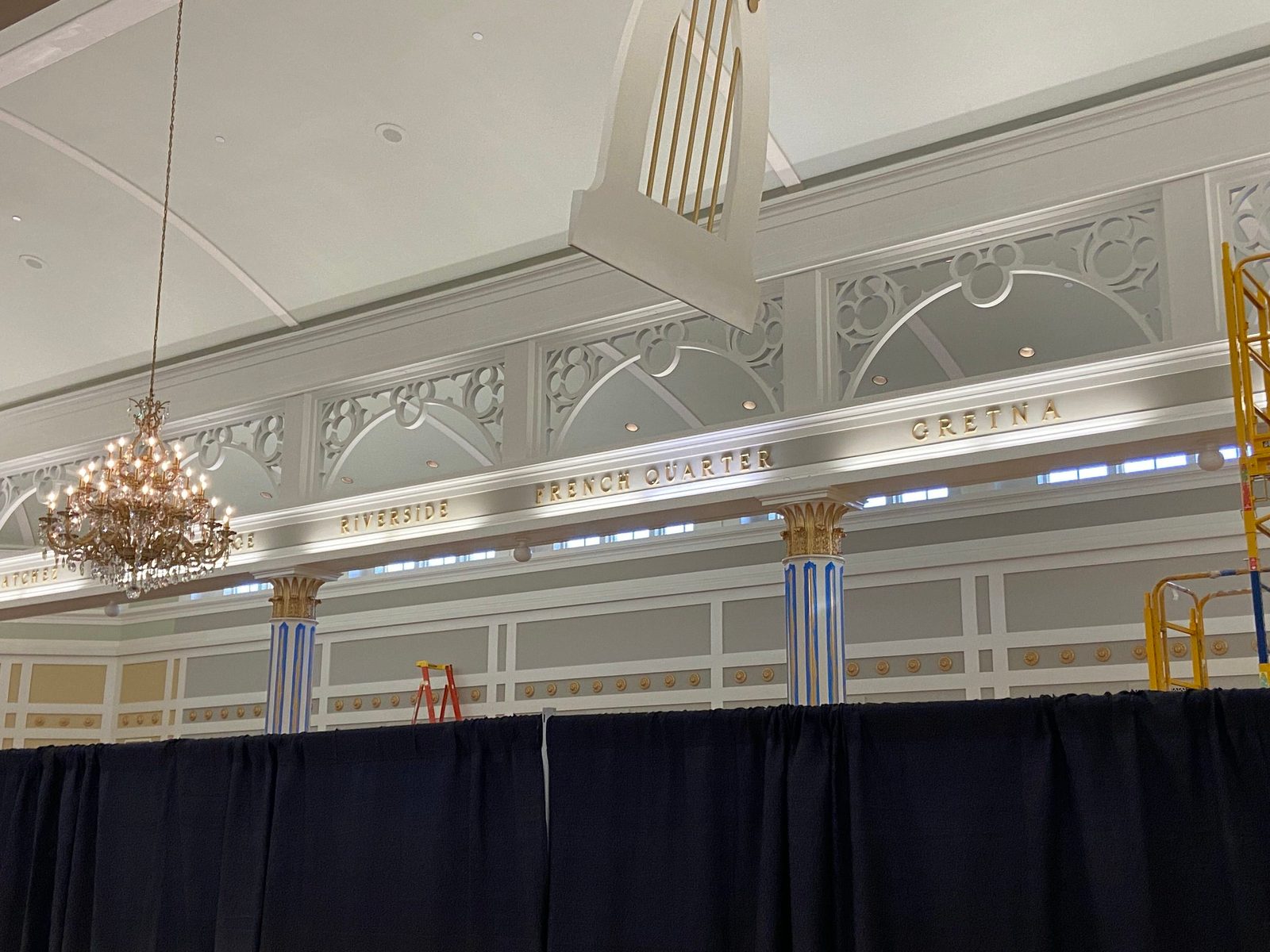 A crystal chandelier hangs above the black curtain line in front of a white wall frieze lettered Riverside, French Quarter and Gretna, with gold-striped columns wrapped in blue painter's tape below and a yellow lift and red ladder poking above the curtains at right