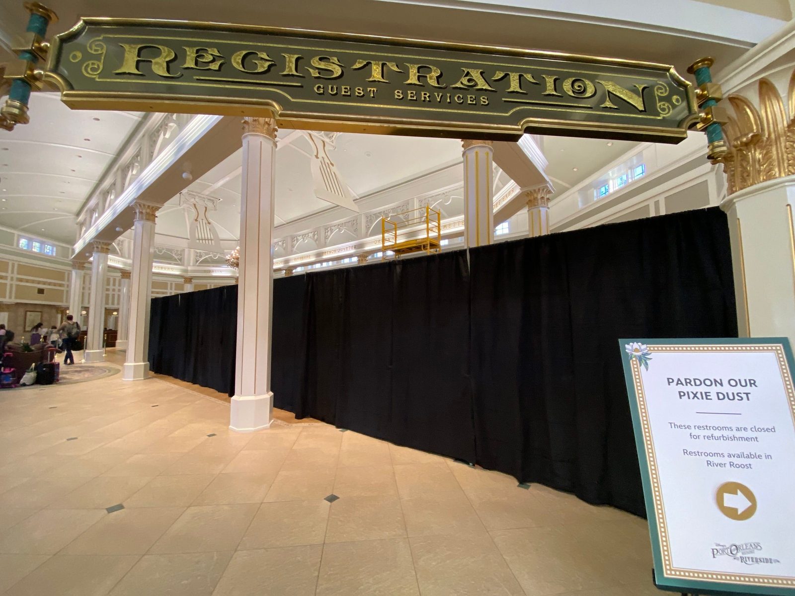 A dark green Registration Guest Services sign with gold lettering hangs above a long row of black curtains in a cream-colored lobby with white columns, while a Pardon Our Pixie Dust easel sign stands in the foreground and a few guests with luggage gather in a seating area at left