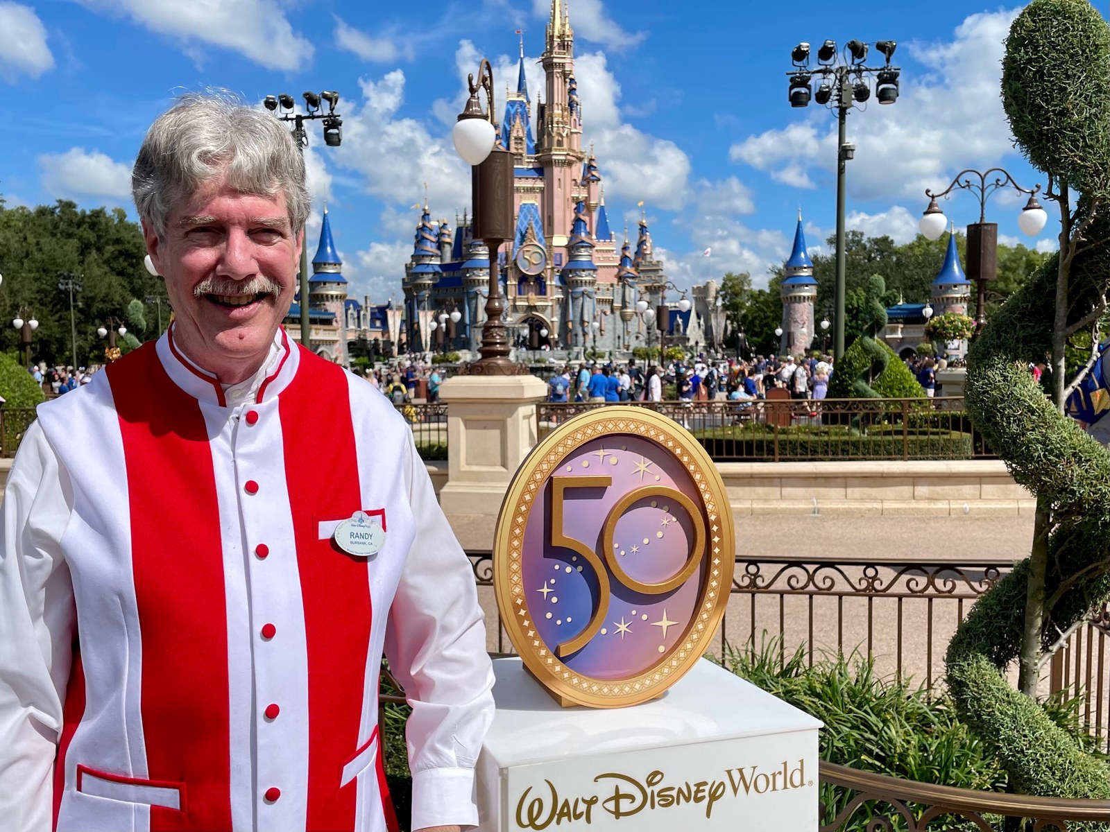 Randy Morris, wearing a red and white striped performer vest and a RANDY name tag, smiles beside a gold 50 anniversary emblem on a Walt Disney World pedestal, with Cinderella Castle and crowds in the hub behind him