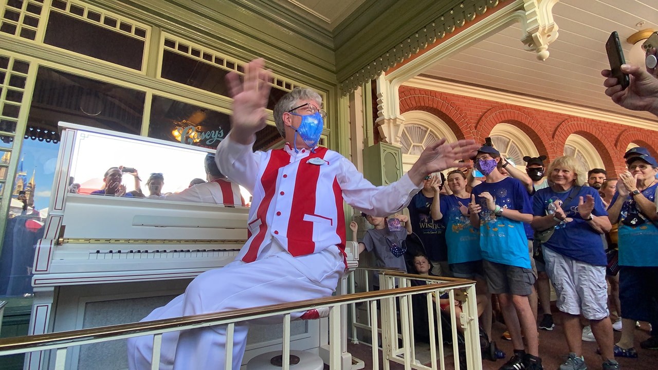 Randy Morris in a red and white striped performer costume and face mask spreads both arms wide from the bench of a white upright piano outside Casey's Corner as a crowd of guests watches and takes photos