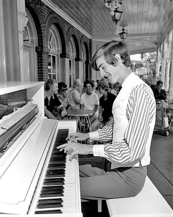 A black and white photo of a young Randy Morris smiling as he plays an upright piano on a covered porch, with guests seated at tables behind him