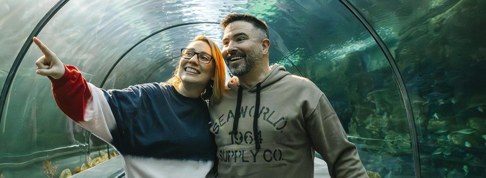A smiling woman points at something overhead while a man in a SeaWorld 1964 Supply Co. hoodie looks on, both standing inside a curved acrylic underwater tunnel with fish swimming around them