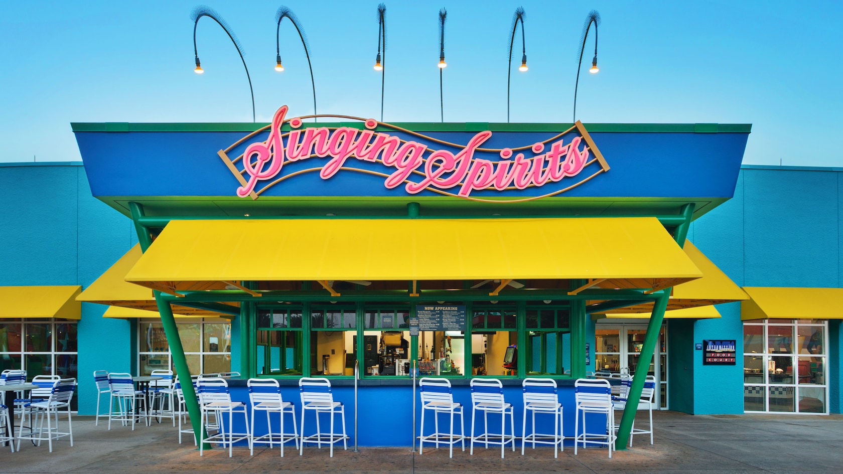 A walk-up bar with a bright blue roof panel and pink neon script sign reading Singing Spirits, a yellow awning over green-framed service windows, and a row of white and blue striped bar stools along the blue counter, with an Intermission Food Court sign on the teal wall to the right