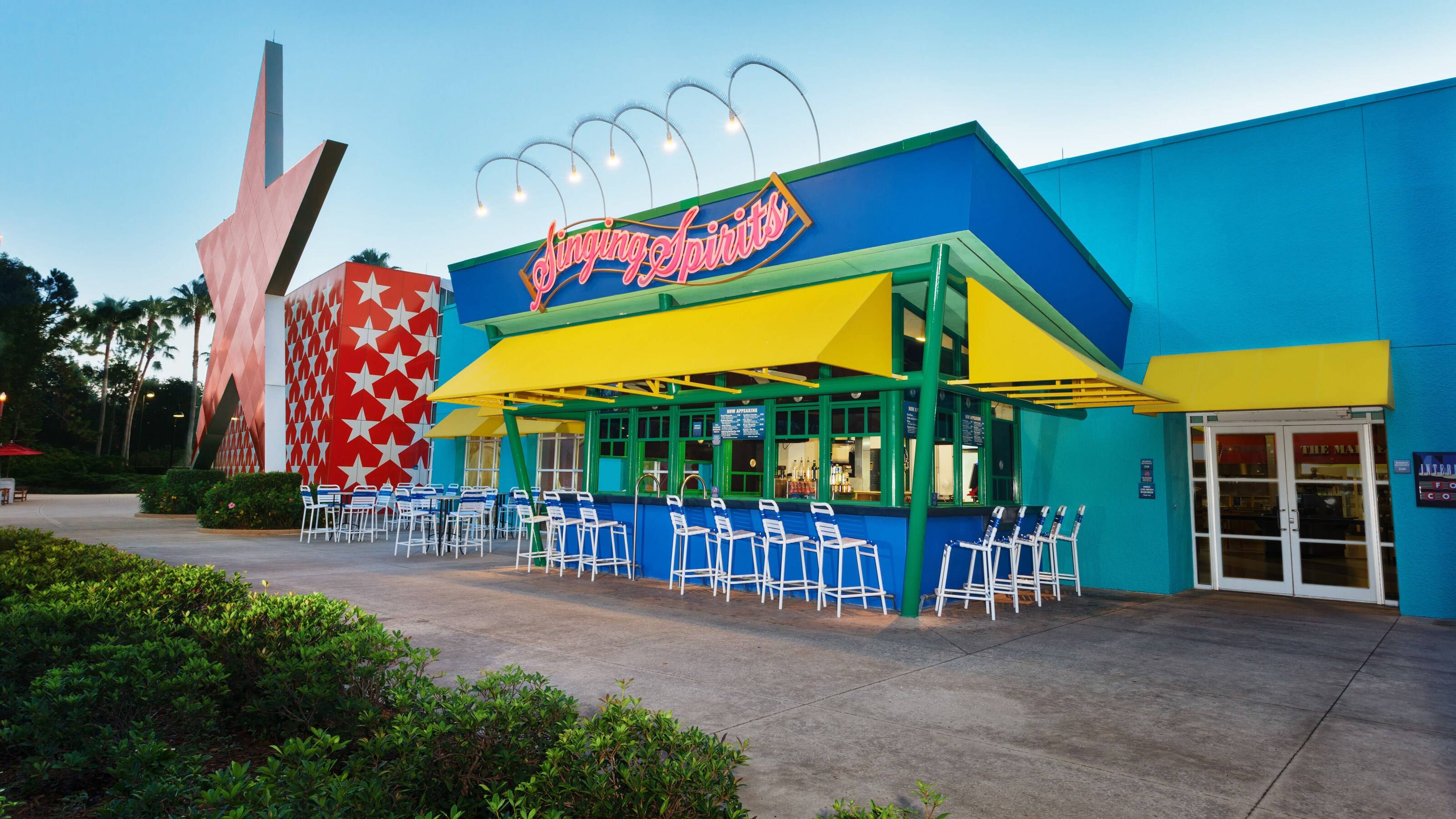 Angled view of the Singing Spirits Pool Bar with its pink neon sign, yellow awnings and rows of white and blue bar stools, next to a giant red star sculpture and a red wall patterned with white stars, with green shrubs and a concrete walkway in the foreground and palm trees behind