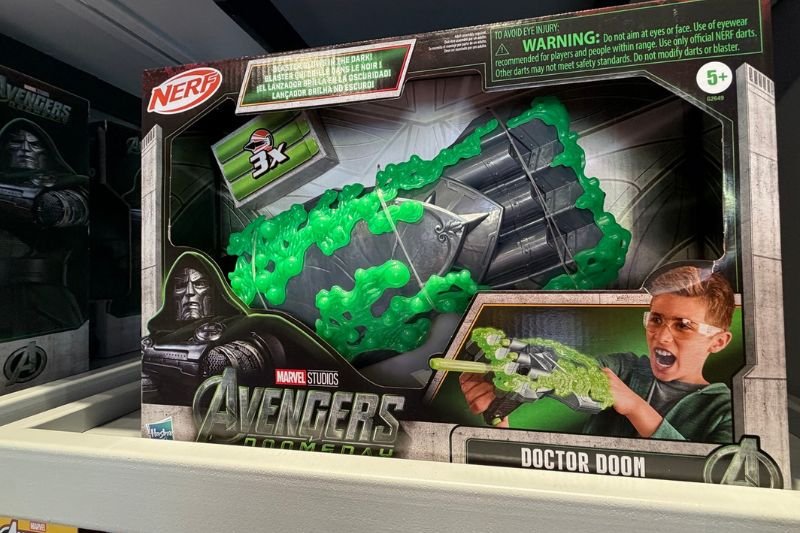 A boxed Nerf blaster on a store shelf, with a gray gauntlet-style blaster wrapped in green, a 3x dart badge, a boy aiming the blaster on the box art, and Marvel Studios Avengers Doomsday and Doctor Doom branding along the bottom