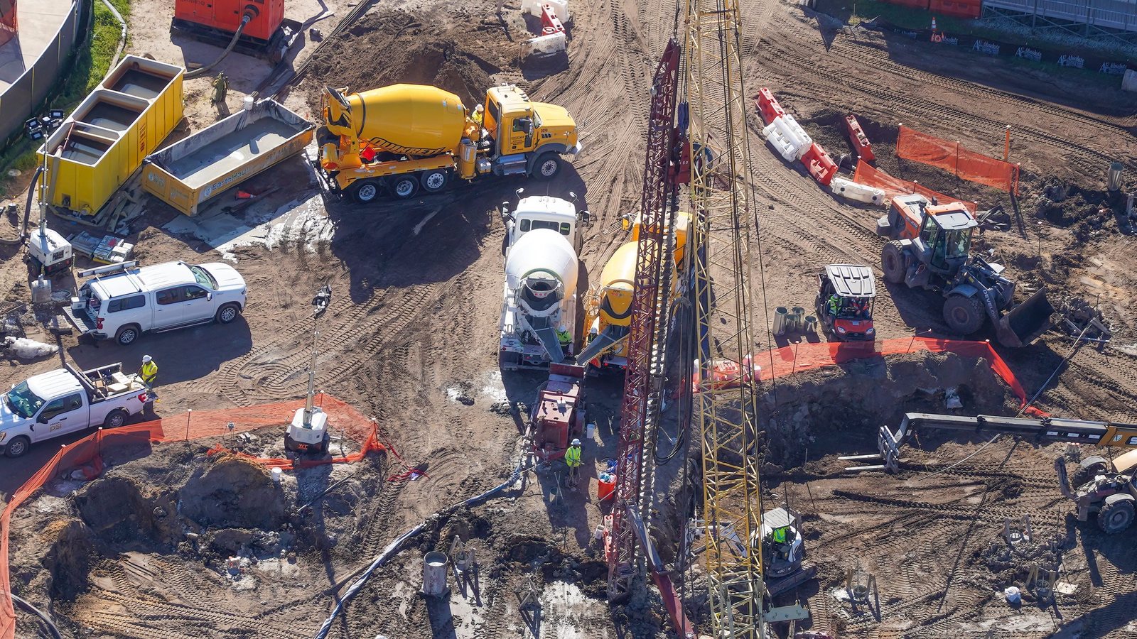 Close aerial view of yellow and white concrete mixer trucks parked beside a tall red and yellow piling rig, with workers in high-visibility clothing, orange safety fencing, a front loader and pickup trucks around them