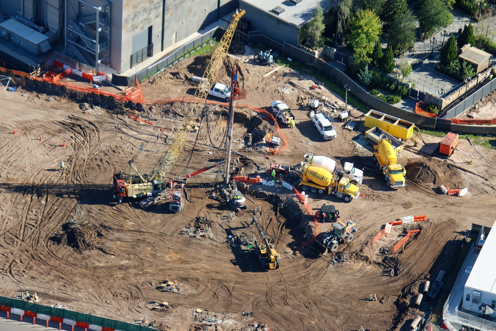 Aerial view of a dirt construction site next to the stone-textured wall of a large show building, with a tall yellow lattice crane, concrete mixer trucks, excavators and orange safety fencing around a trench