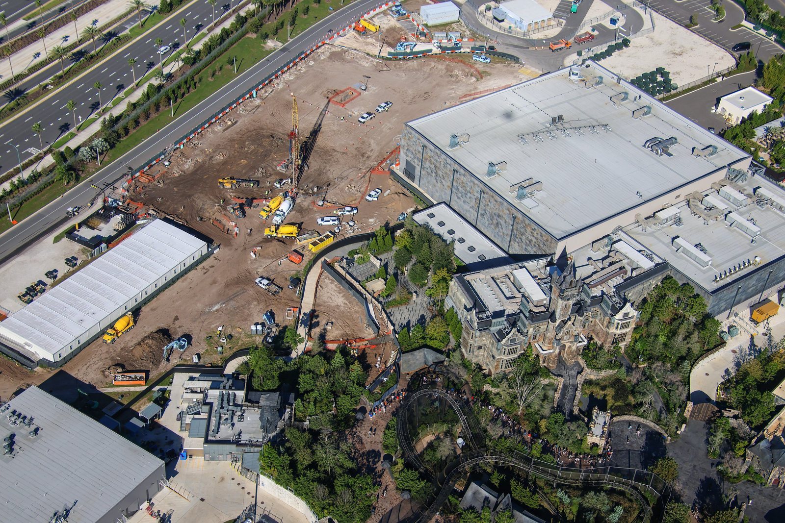 Aerial view of a large dirt construction site with a tall crane-mounted rig, concrete mixer trucks and white pickups, bordered by a multi-lane road on the left and a large gray show building and a Gothic manor on the right, with a roller coaster track in trees at the bottom