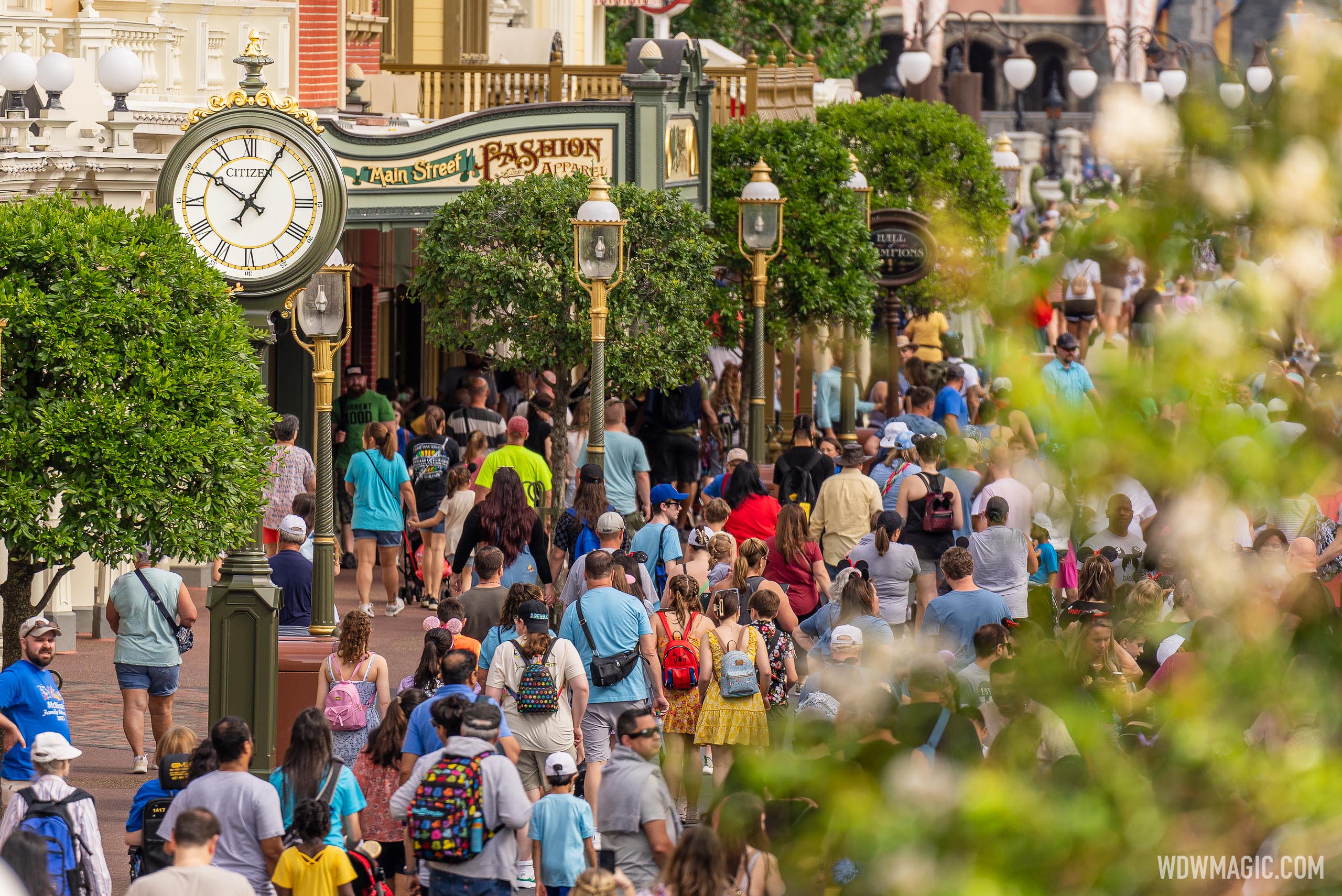 Crowds of guests walking up Main Street, U.S.A. at Magic Kingdom past a green Citizen street clock and the Main Street Fashion and Apparel sign, with Cinderella Castle partly visible in the background