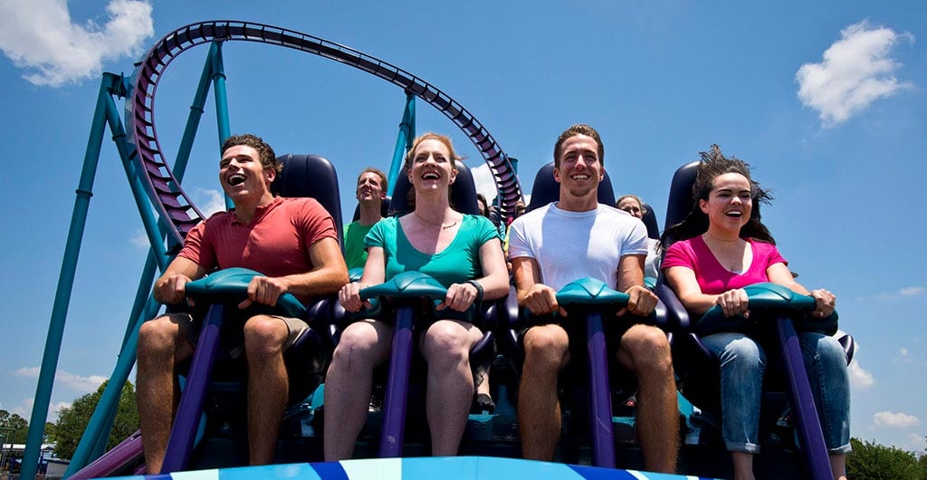 Four smiling riders in Mako's front row, each held by a teal-topped lap restraint mounted on a purple bar between their knees, with Mako's purple track and teal supports arcing against a blue sky behind them