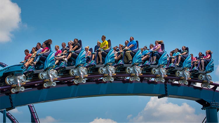 A Mako train of riders cresting a hill on blue and purple track against a blue sky, each rider seated in an open row with a lap restraint