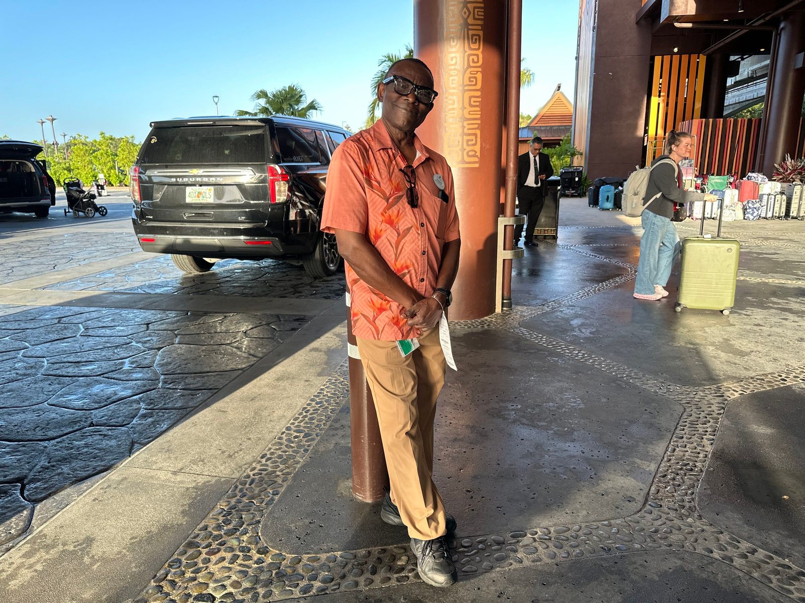 A bellhop in an orange button-up shirt with a bird-of-paradise flower print on one half and khaki pants standing at the resort's drop-off area, with a black SUV and a guest with luggage behind him